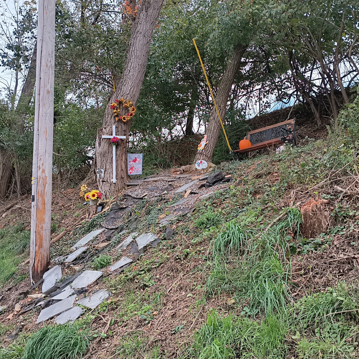 Roadside memorial for a car accident victim on an embankment. There's a bench at the top of the embankment with a pumpkin, a cross and wreath nailed to a tree, a gnome mailed to another tree, a lawn flag with cardinals on it, some plants, and paver stones up the side of the embankment.