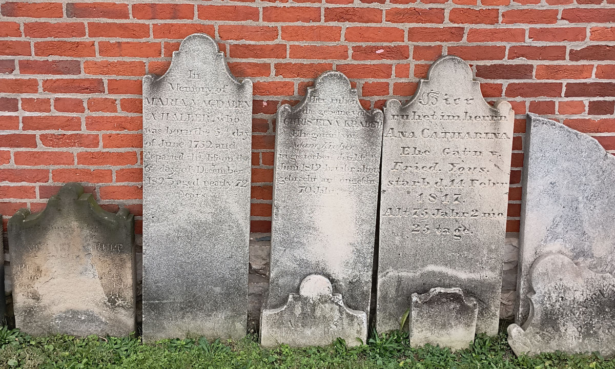 Close up of three of the headstones against the wall. The one on the left is in English; the center and right headstones are in German.